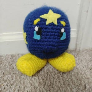 May include: A handmade crochet plush toy in the shape of a blue sphere with yellow star and crescent accents. The toy has yellow feet and features a face with blue tears and a smiling mouth. The toy is made of yarn.