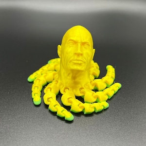 May include: A yellow 3D printed sculpture of a human head with a face, sitting on top of a yellow octopus with green tentacles.