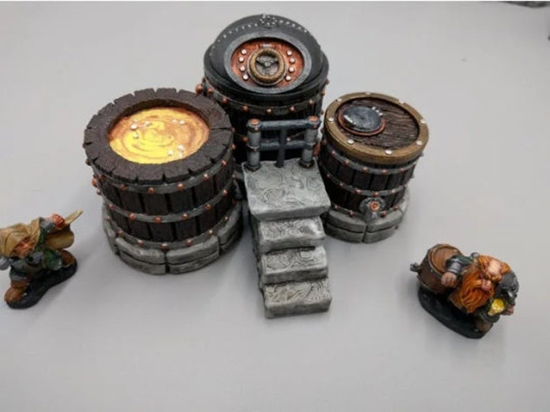 May include: A miniature set of three wooden barrels with metal accents, one with a golden liquid inside, and two dwarves figurines. The barrels are on a stone platform with steps leading up to a metal railing.