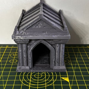 May include: A grey, 3D printed miniature mausoleum with a detailed roof and arched doorway. The mausoleum is sitting on a green cutting mat with yellow lines.