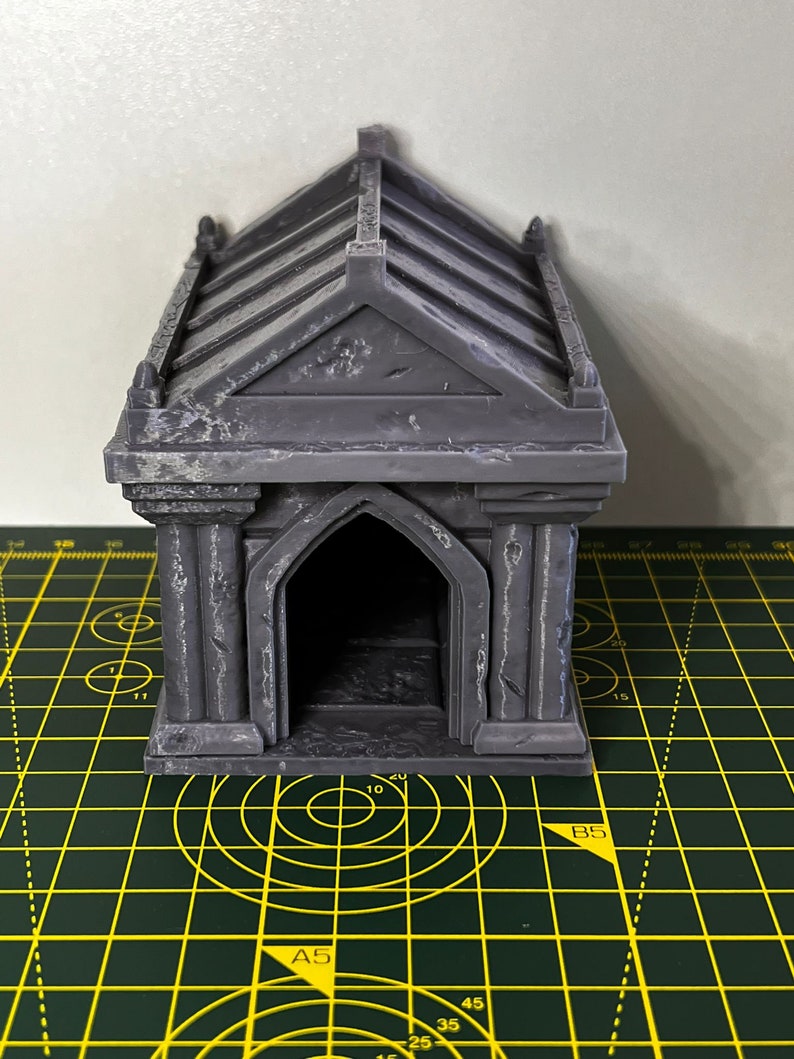 May include: A grey, 3D printed miniature mausoleum with a detailed roof and arched doorway. The mausoleum is sitting on a green cutting mat with yellow lines.