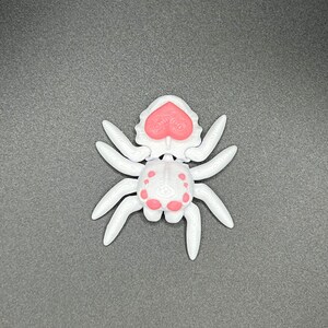 May include: A white plastic spider toy with pink accents. The spider has a heart-shaped head and eight legs.