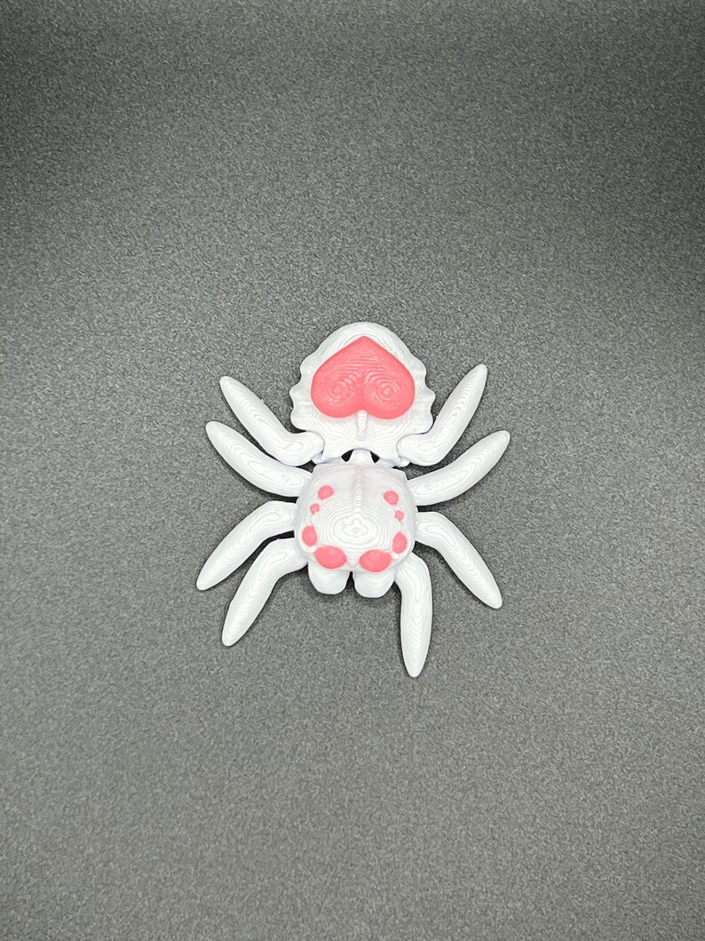 May include: A white plastic spider toy with pink accents. The spider has a heart-shaped head and eight legs.