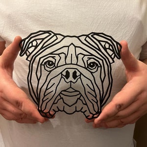 May include: A white t-shirt featuring a geometric line art design of a bulldog's face. The black lines create a striking contrast against the white fabric. The design is centered on the shirt.