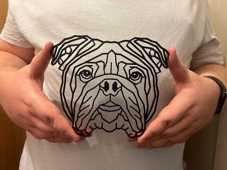 May include: A white t-shirt featuring a geometric line art design of a bulldog's face. The black lines create a striking contrast against the white fabric. The design is centered on the shirt.