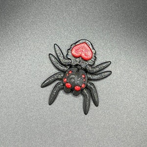 May include: A black spider with red heart-shaped eyes and red dots on its body. The spider is made of plastic and has a 3D printed look.