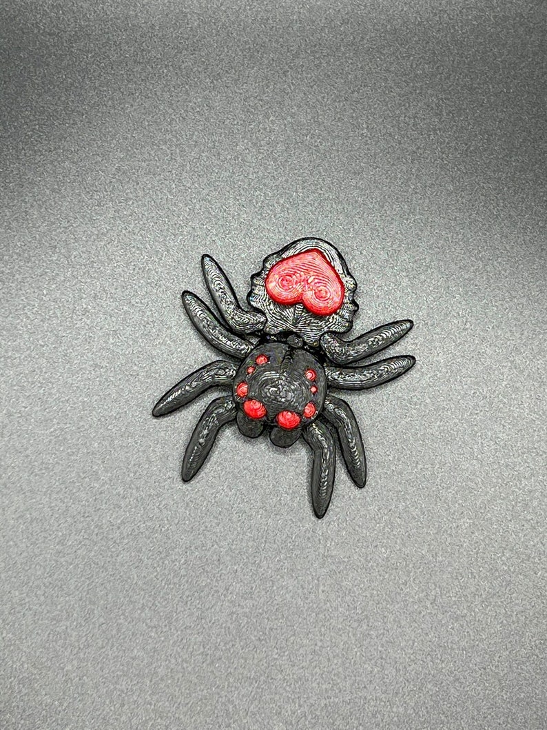 May include: A black spider with red heart-shaped eyes and red dots on its body. The spider is made of plastic and has a 3D printed look.