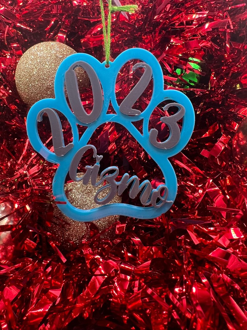 May include: Blue paw print Christmas ornament with the year 2023 and the name Gizmo written inside.