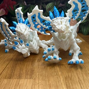 Puede incluir: Dos figuras de drag&oacute;n blancas impresas en 3D con detalles azules. Los dragones tienen alas, garras y espinas detalladas.