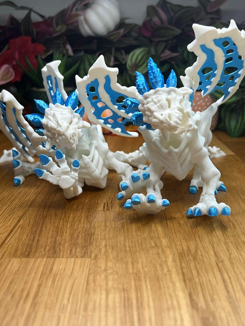 Puede incluir: Dos figuras de drag&oacute;n blancas impresas en 3D con detalles azules. Los dragones tienen alas, garras y espinas detalladas.