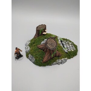 May include: A miniature terrain piece featuring a pair of tree stumps with moss and a stone path. The piece is designed for tabletop gaming.