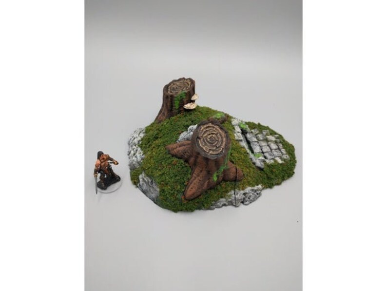 May include: A miniature terrain piece featuring a pair of tree stumps with moss and a stone path. The piece is designed for tabletop gaming.