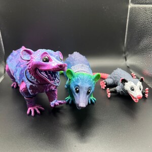 May include: Three small opossum figurines, one is pink and purple with a white face, one is blue and green with a white face, and one is gray with a white face and pink ears.