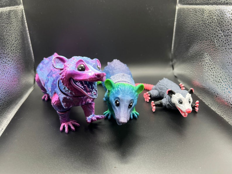 May include: Three small opossum figurines, one is pink and purple with a white face, one is blue and green with a white face, and one is gray with a white face and pink ears.