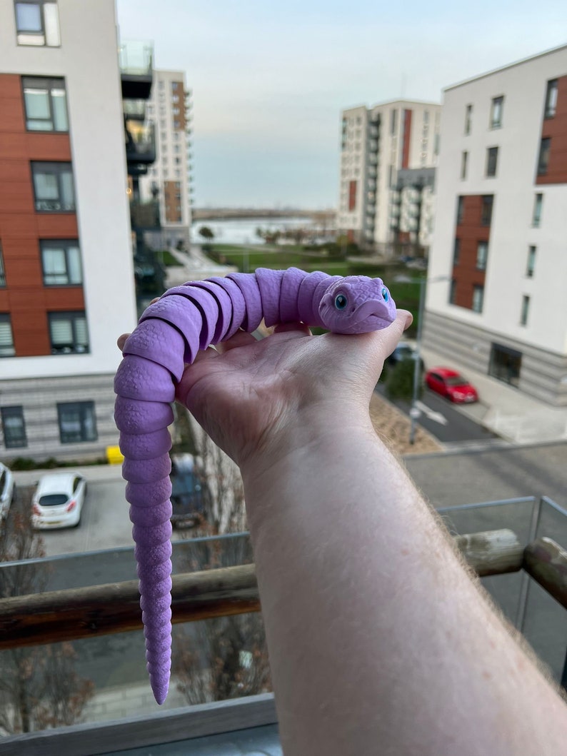 May include: A purple, segmented, bendable toy snake with a face and blue eyes. The snake is being held by a hand in front of a city skyline.