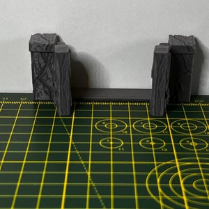 May include: Two gray, wooden, textured, miniature gate pieces on a green and yellow grid surface.