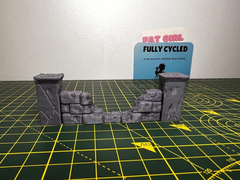 May include: A gray, 3D printed, miniature stone wall with a broken section in the middle. The wall is made of rough, textured blocks. The wall is on a green cutting mat with white markings.
