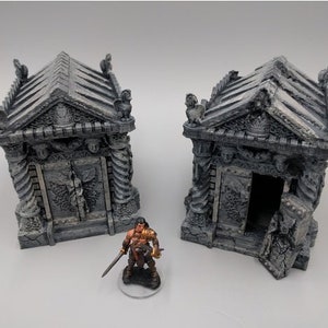 May include: Two gray stone mausoleums with intricate details and open doorways. A miniature figure of a warrior stands in front of the mausoleums.