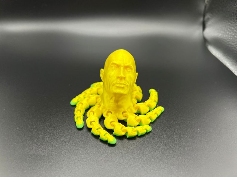 May include: A yellow 3D printed sculpture of a human head with a face, sitting on top of a yellow octopus with green tentacles.