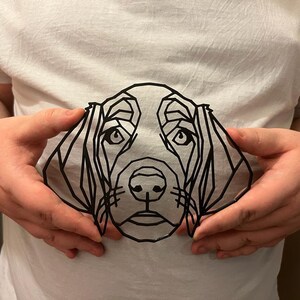 Op de afbeelding: Een wit t-shirt met een geometrische zwarte lijntekening van een hondengezicht. De kenmerken van de hond worden gedefinieerd door hoekige lijnen, waardoor een modern, minimalistisch ontwerp ontstaat. Het shirt wordt door een persoon vastgehouden.