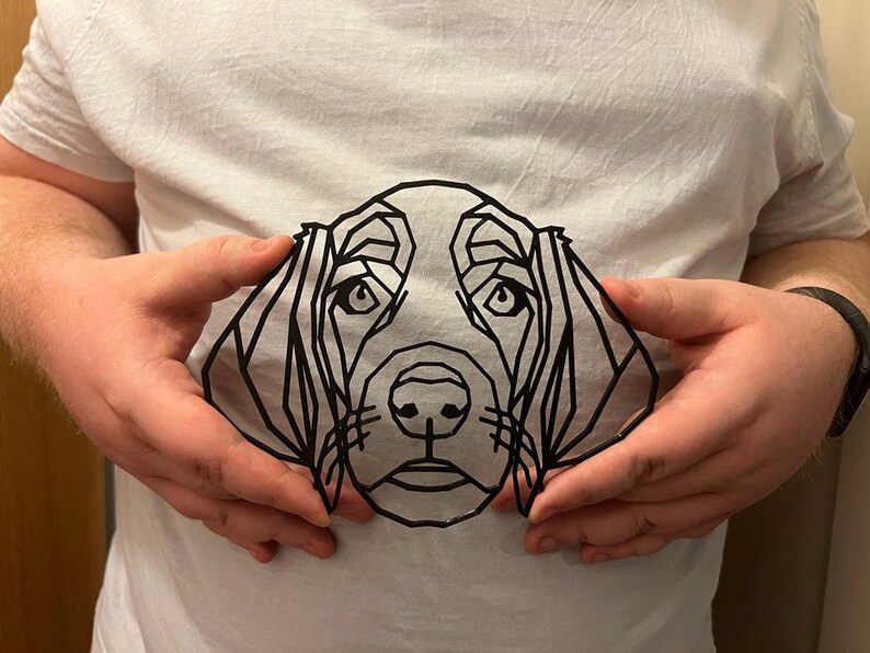 Op de afbeelding: Een wit t-shirt met een geometrische zwarte lijntekening van een hondengezicht. De kenmerken van de hond worden gedefinieerd door hoekige lijnen, waardoor een modern, minimalistisch ontwerp ontstaat. Het shirt wordt door een persoon vastgehouden.