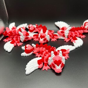 Puede incluir: Cinco peque&ntilde;os dragones alados de color rosa y blanco con corazones rojos en sus cuerpos. Los dragones est&aacute;n acostados sobre una superficie negra.