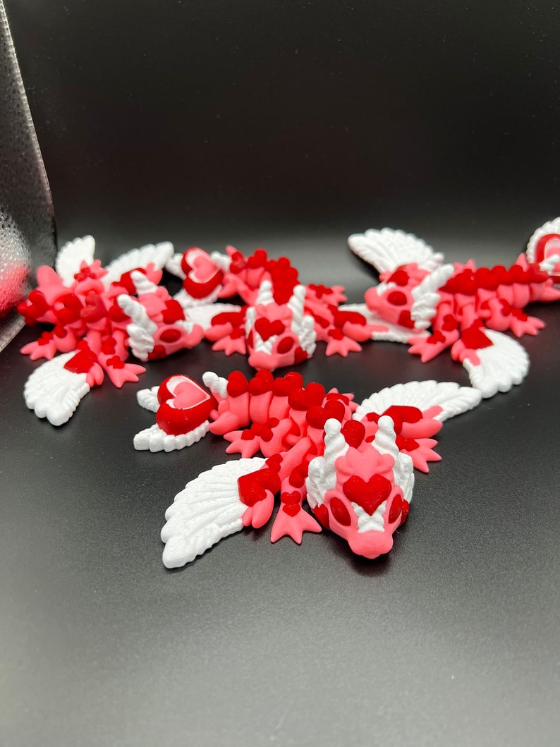 Puede incluir: Cinco peque&ntilde;os dragones alados de color rosa y blanco con corazones rojos en sus cuerpos. Los dragones est&aacute;n acostados sobre una superficie negra.