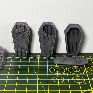 May include: Three gray plastic miniature coffins. One coffin is closed, one is open with a broken lid, and one has a chain and padlock on the lid.