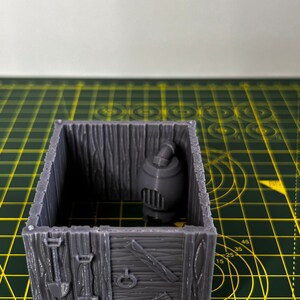May include: A gray 3D printed miniature wooden shack with a small gray stove inside. The shack has a wooden door and a window. The shack is sitting on a green cutting mat.