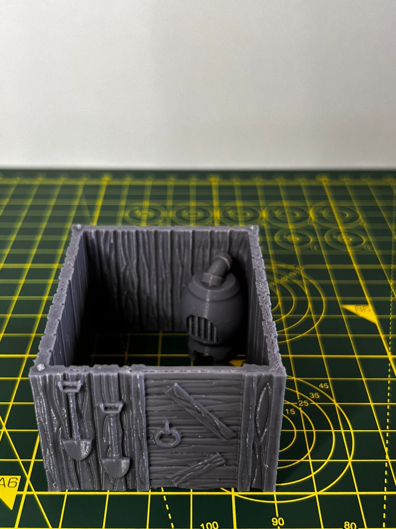 May include: A gray 3D printed miniature wooden shack with a small gray stove inside. The shack has a wooden door and a window. The shack is sitting on a green cutting mat.