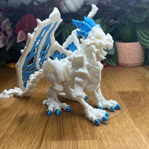 Puede incluir: Esqueleto de drag&oacute;n articulado blanco con detalles azules. El drag&oacute;n tiene una cola larga, alas y garras. El drag&oacute;n est&aacute; en posici&oacute;n de pie con la cabeza girada hacia un lado.