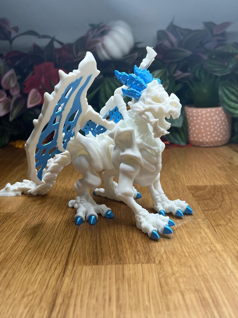 Puede incluir: Esqueleto de drag&oacute;n articulado blanco con detalles azules. El drag&oacute;n tiene una cola larga, alas y garras. El drag&oacute;n est&aacute; en posici&oacute;n de pie con la cabeza girada hacia un lado.