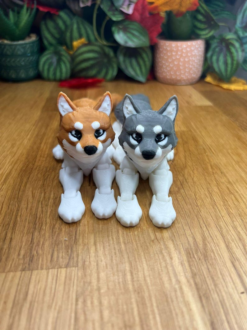 Peut inclure: Deux petites figurines de loup articul&eacute;es imprim&eacute;es en 3D. Le loup &agrave; gauche est orange et blanc, et le loup &agrave; droite est gris et blanc. Les deux loups sont couch&eacute;s, les pattes tendues.