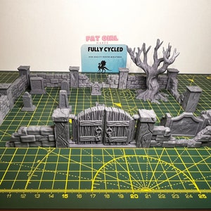 May include: A 3D printed miniature graveyard set with a gate, walls, tombstones, and a bare tree. The set is made of gray plastic and is designed for tabletop gaming.