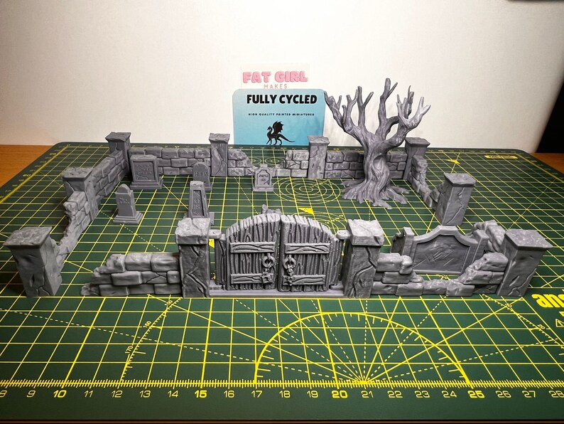 May include: A 3D printed miniature graveyard set with a gate, walls, tombstones, and a bare tree. The set is made of gray plastic and is designed for tabletop gaming.