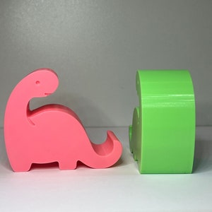 May include: Two 3D printed dinosaur phone holders, one pink and one green. The pink dinosaur is facing left and the green dinosaur is facing right.