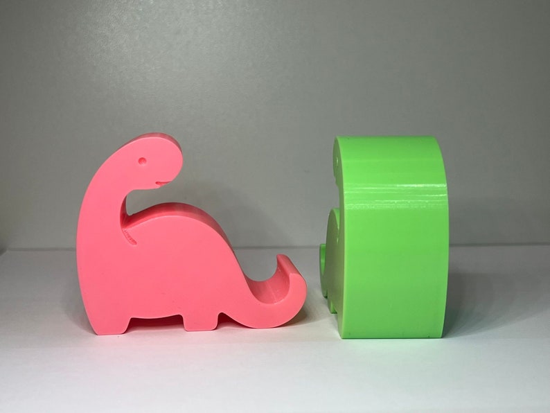 May include: Two 3D printed dinosaur phone holders, one pink and one green. The pink dinosaur is facing left and the green dinosaur is facing right.