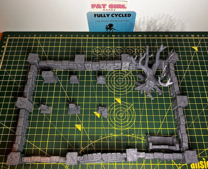 May include: A set of gray 3D printed miniature dungeon walls and a tree with roots, on a green cutting mat. The walls are made of stone and have a rough texture. The tree is made of a smooth material and has a realistic appearance. The text on the card reads "Fat Girl Makes Fully Cycled High Quality Printed Miniatures".