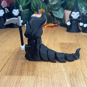May include: Black skeletal figure with a scythe and a segmented tail. The figure is wearing a black hooded robe and has a white face.