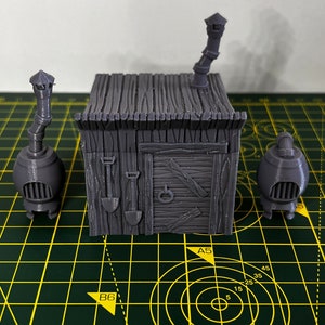 May include: A 3D printed gray model of a small wooden house with a chimney and a door. The house is sitting on a green cutting mat with yellow lines.