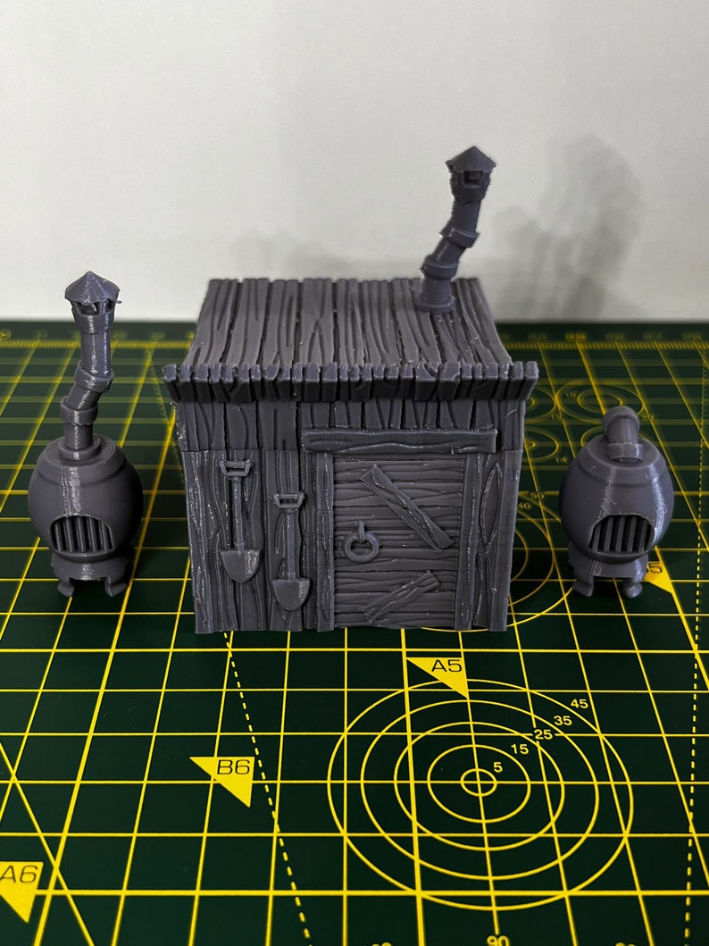May include: A 3D printed gray model of a small wooden house with a chimney and a door. The house is sitting on a green cutting mat with yellow lines.