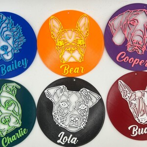 May include: Six round, colorful ornaments with geometric dog illustrations and names. The ornaments are blue, orange, purple, green, black, and red. The names are Bailey, Bear, Cooper, Charlie, Lola, and Buddy.