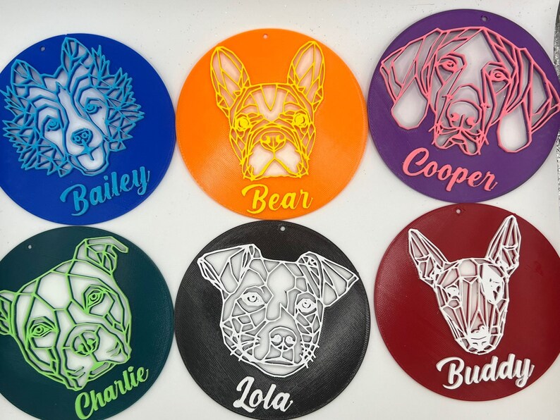 May include: Six round, colorful ornaments with geometric dog illustrations and names. The ornaments are blue, orange, purple, green, black, and red. The names are Bailey, Bear, Cooper, Charlie, Lola, and Buddy.