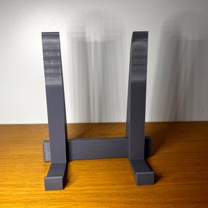 May include: A gray plastic stand with two vertical supports and a horizontal base. The stand is designed to hold a plate or other flat object.