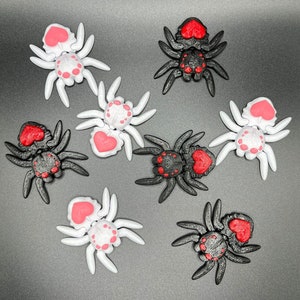 May include: Eight small plastic spiders with red hearts on their backs. Four spiders are black and four spiders are white.