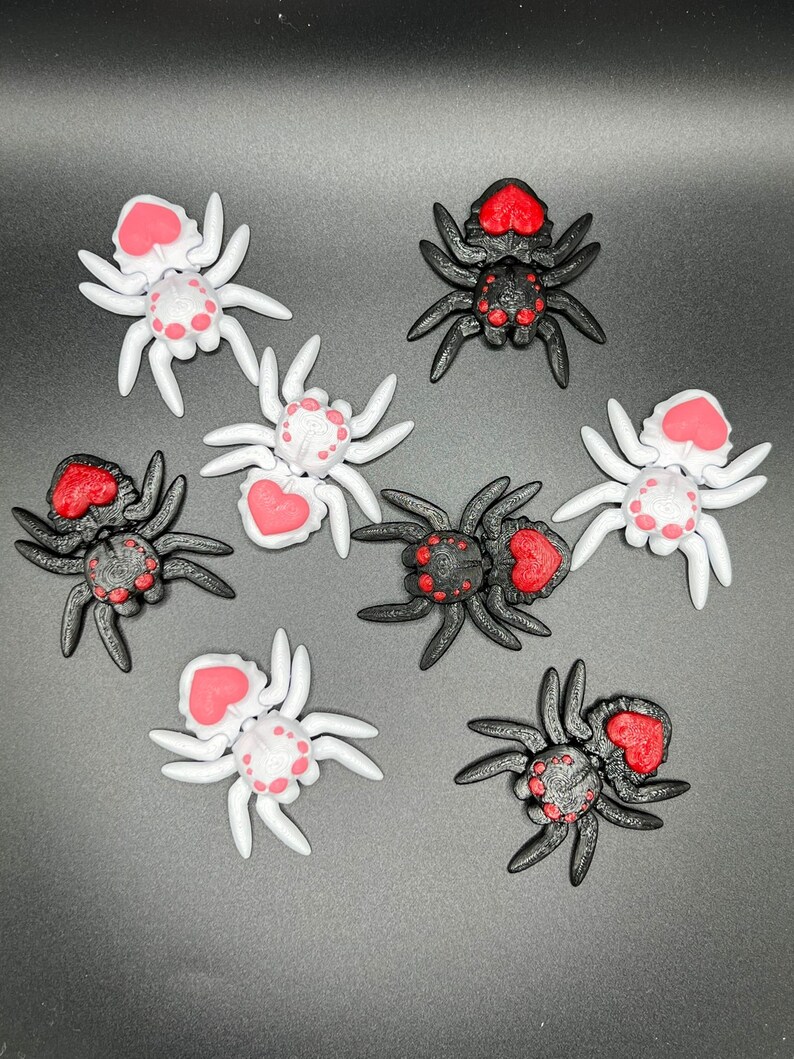 May include: Eight small plastic spiders with red hearts on their backs. Four spiders are black and four spiders are white.