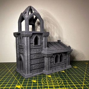 May include: A gray 3D printed miniature of a gothic church with a bell tower. The church has a stone facade with arched windows and a pointed roof.