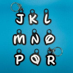 Op de afbeelding: Negen zwarte sleutelhangers met witte letters die het woord "JKLMNOPQR" spellen op een blauwe achtergrond.