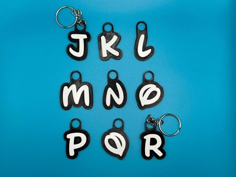 Op de afbeelding: Negen zwarte sleutelhangers met witte letters die het woord "JKLMNOPQR" spellen op een blauwe achtergrond.