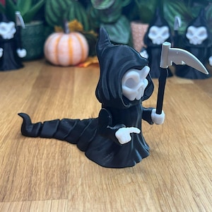 May include: A black and white 3D printed figurine of a skeletal figure in a hooded robe, holding a scythe. The figure is in a crawling pose with a long, segmented tail.